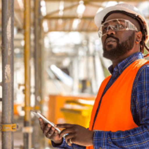 A man wearing an orange vest and safety glasses stands in front of a construction site, overseeing the work.